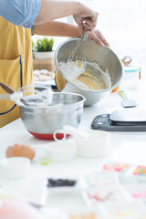 Hands of woman with apron mixing cake batter for cupcakes with whisk in kitchen at home. Spatula,...