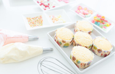 Selective focus on fresh baked cupcakes. Piping bags with pastel butter cream, icing candy, chocolate flakes or coated sugar and jelly in tray for decorating. egg whisk on white table