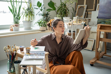 Inspired female artist drawing with watercolor in sketchbook, checking textbook on working with paints. Focused woman learning studying, practicing skill of painting by reading book, manual, guide.