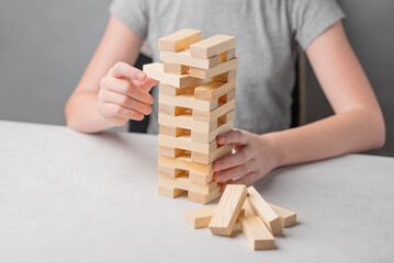 A child sits at a table and with his hand pulls out a piece from a wooden tower game on a light...