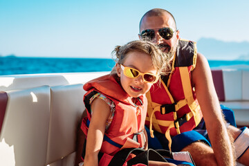 A little girl and her father, both wearing orange life jackets, seated on a boat, enjoying a...