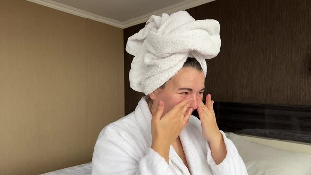 Horizontal 4K video. White woman in towel on head, robe applies moisturizer cream to face in hotel bed room. Skin protection during cold season. Spa salon. Rest, relax, march 8, body care, 14 february