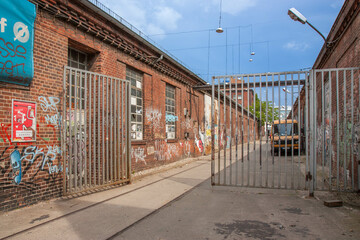  An old alley with old buildings built of red brown bricks, broken windows and graffiti. A gray rusty open gate