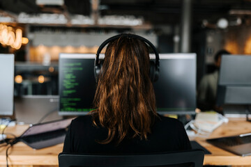 Rear view of female coder with headphones working on computer at office