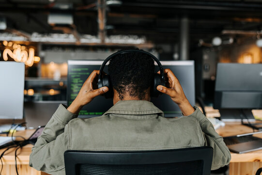 Rear view of male coder wearing wireless headphones sitting at desk in office