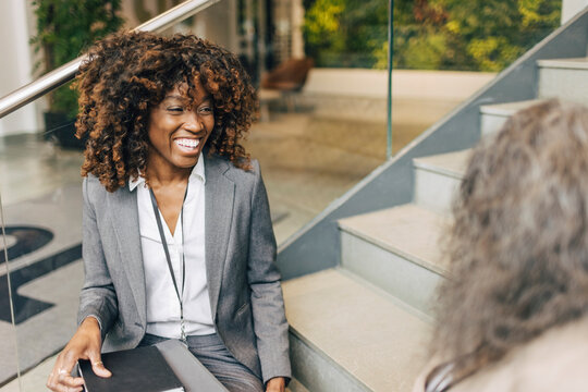 Happy female entrepreneur with curly hair sitting on steps while talking to delegate at convention center
