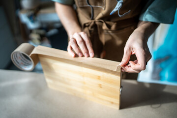 Hands of female artist carefully prepare wooden board for working with epoxy resin, glue edges with paper tape. Close-up, woman processes canvas for future painting artwork in technique of liquid art