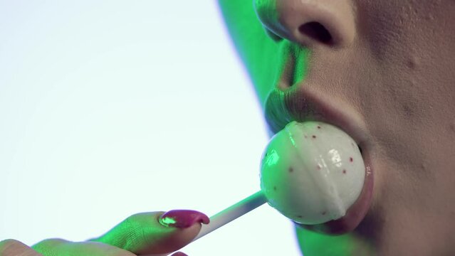 Kobieta na białym tle bierze do ust kolorowy lizak w kropki i powoli go oblizuje. Jej ponętne, nawilżone usta błyszczą, a z tyłu twarz oświetla zielone światło. Makro, zmysłowy detal