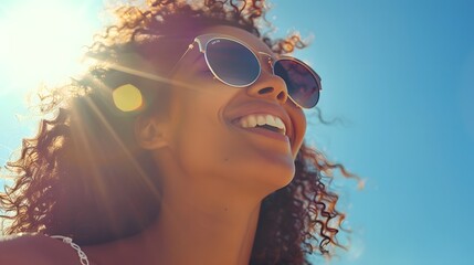 A woman wearing sunglasses smiles as she looks up at the sun on an sunny day, with blue sky in background. She is standing outdoors and has long curly hair.