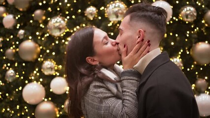 Cheerful romantic couple kissing in front of decorated Christmas tree with golden baubles. Side view of affectionate man and woman spending quality time during festive season. 