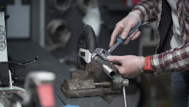Professional mechanic works in garage, uses bench vise to clamp spare part from scooter and repair. Repairman fixes wheel from scooter clamped in cradle and twisting with crescent wrench in workshop. 