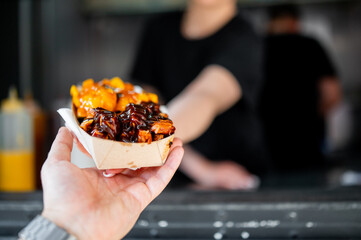 Close-up of a hand holding a paper tray with delicious-looking cheesy fries topped with a rich,...