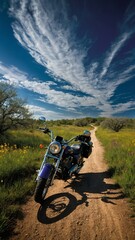 Motorcycle stands on dirt road, cutting through field adorned with wildflowers. Road leads towards horizon under sky filled with wispy clouds. Shadow of motorcycle cast on ground.