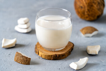 A glass of fresh coconut milk placed on a wooden coaster, surrounded by coconut pieces on a textured gray surface.