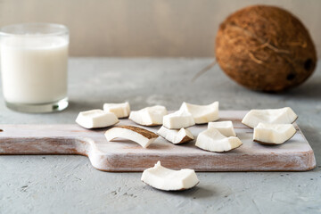 Sliced coconut pieces arranged on a wooden cutting board with a glass of coconut milk and a whole coconut in the background.