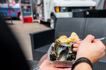 Hand holding a fork, enjoying food from a paper container with food trucks in the background