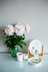 Bouquet of pink peonies in glass vase, candles, cosmetics and women's things on wooden chest of drawers on white background. Spring flowers.