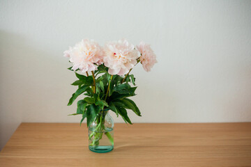 Bouquet of pink peonies in glass vase on wooden chest of drawers on white background. Spring flowers.