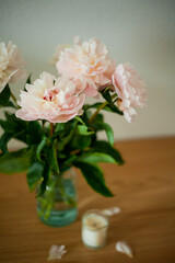 Bouquet of pink peonies in glass vase, candles on wooden chest of drawers on white background. Spring flowers.