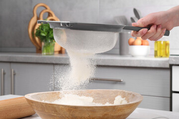 Woman sieving flour into bowl at white wooden table in kitchen, closeup