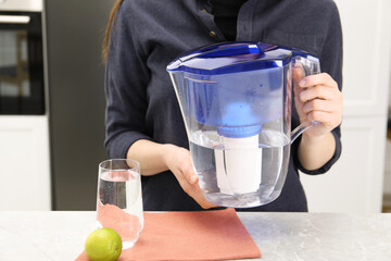Woman with water filter jug at light marble table in kitchen, closeup