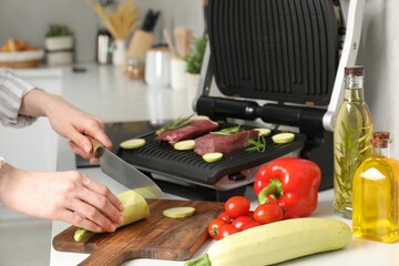 Woman cooking different products with electric grill at white wooden table in kitchen, closeup