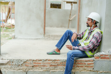 Asian male construction worker in reflective vest and hard hat sitting on unfinished building structure. looks thoughtful and tired, taking break from work. construction site and equipment.
