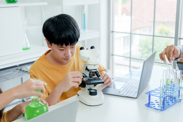 Students' learning in science classroom. Focused young boy students experiment with chemicals, using flasks and test tubes, with microscope and laptop on clean white desk in classroom.