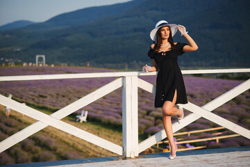 Sexy woman in elegant black dress and hat posing in bloomig lavender field. Fashion outdoor photo of beautiful girl.