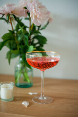 Bouquet of pink peonies in glass vase, candles, glass with a pink cocktail on wooden chest of drawers on white background. Spring flowers.