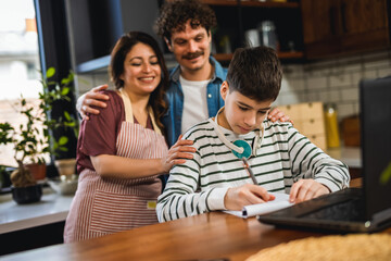Latino boy doing his homework with the help of computer. Proud parents are watching him.