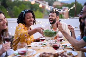 Smiling African American woman sitting at table passing salad to happy friends at meal event outdoors. Multiracial cheerful young people enjoying BBQ reunion together at weekend on rooftop at dusk