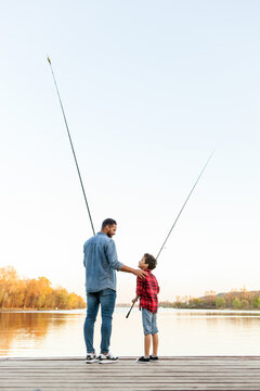 African American man and child standing on wooden pier holding fishing rods on the river, dad teaching his son to fish in the lake, family resting and relaxing on the weekend
