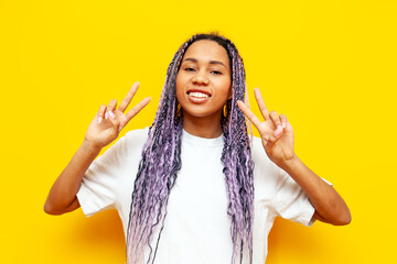cheerful african american woman with colored dreadlocks showing peace gesture on yellow isolated...