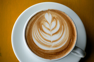 A detailed close-up of latte with intricate leaf art in white cup, set on yellow wooden table