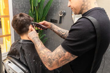   tattooed barber cutting the hair and beard of a young, dark-haired client.