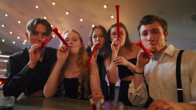 People celebrating b-day, blowing noisemakers whistles, congratulation party. Portrait of happy five best friends cheering and greeting, merry birthday party, gathering in festival in nightclub