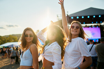 Three happy female friends dance at sunny beach music fest. Crowd enjoys summer vibes near stage....