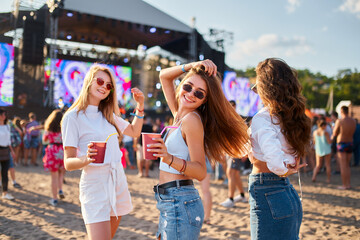Group of joyful girls with drinks dance at beach music fest under summer sun. Happy females enjoy cocktails, vibes near stage with crowd. Friendship, party lifestyle at tropical festival event.