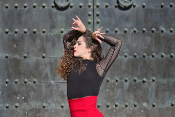 Obraz premium Portrait of young, beautiful, brunette woman in black shirt and red skirt, dancing flamenco with old, black metal door in the background. Flamenco concept, dance, art, typical Spanish.