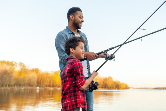 African American man and child standing on wooden pier holding fishing rods on the river, dad teaching his son to fish in the lake, family resting and relaxing on the weekend