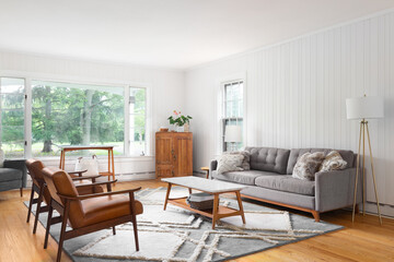 A cozy living room with wooden chairs and a grey sofa, a marble top table on a rug and hardwood floors, and a large bay window looking outside. No brands or labels.