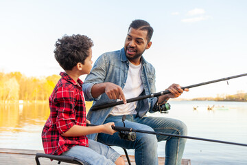 African American man and child sitting on a wooden pier holding fishing rods on the river, dad teaching his son to fish in the lake, family resting and relaxing on the weekend