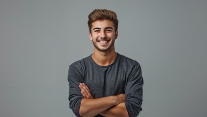 A handsome young man smiling with his arms crossed against a grey background, wearing casual with a confident and happy expression in a studio portrait.