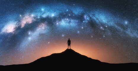 Glowing Milky Way and man on mountain peak at starry night. Silhouette of a guy on the hill, sky with bright stars. Galaxy. Space landscape with milky way arch. Panoramic nightscape. Travel. Concept