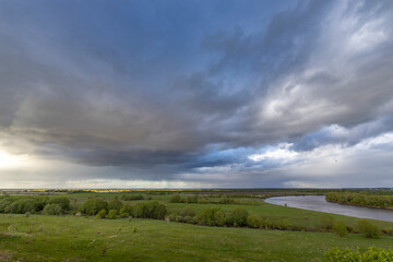 A serene meadow overlooks a winding river under a dramatic sky filled with storm clouds and patches of blue, capturing the beauty of nature's ever-changing weather patterns