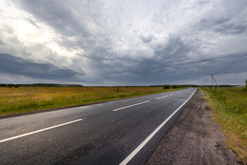 A paved road stretches out through a field, with a vast, gray sky overhead. The air is filled with the anticipation of a storm.