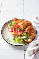 Croissant sandwich with salami sausage, cheese and cucumber in a plate for tasty breakfast on white tile table, text space
