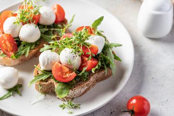 Sandwiches with tomatoes, mozzarella balls, arugula on white plate light textured background close up. Italian bruschetta set, antipasti