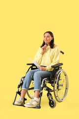 Thoughtful young woman in wheelchair with notebook on yellow background